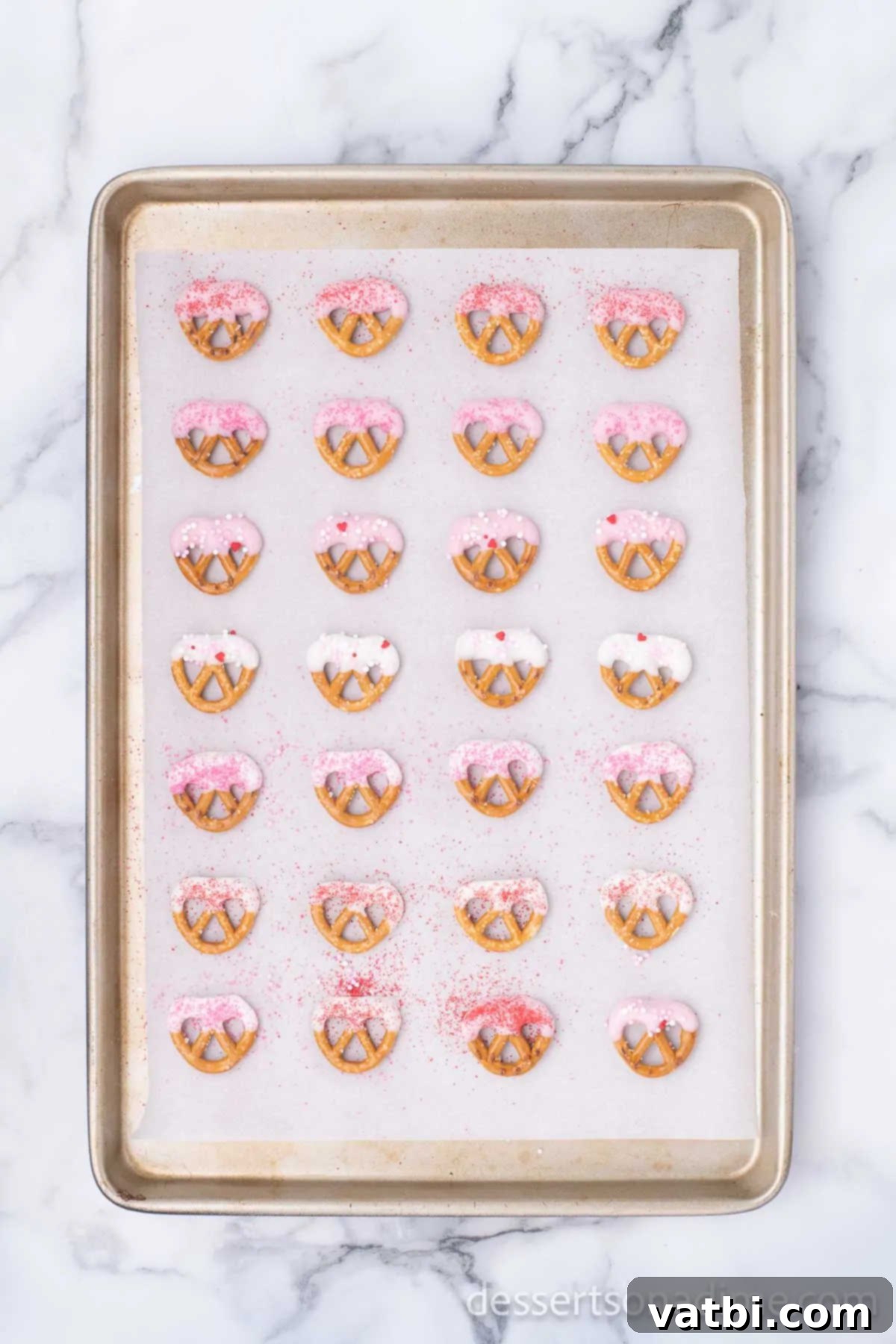 Love Crunch Pretzel Bites 3 A baking sheet covered with parchment paper, displaying several chocolate-covered pretzels drying, adorned with colorful sprinkles.