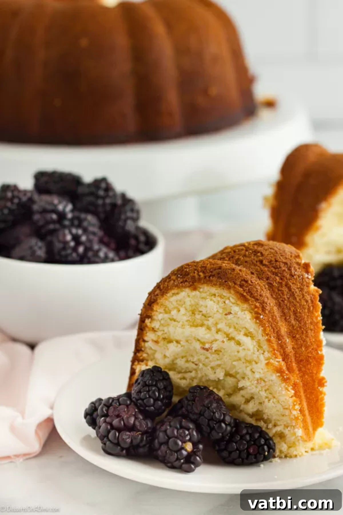 Learn how to make this easy and simple pound cake recipe. How to make moist old fashion pound cake is easier than you think and everyone loves it. This is the best pound cake recipe from scratch! #dessertsonadime #cakerecipes #dessertrecipes A second close-up image of a slice of pound cake on a white plate, adorned with fresh blackberries, emphasizing its golden crust and fluffy texture.