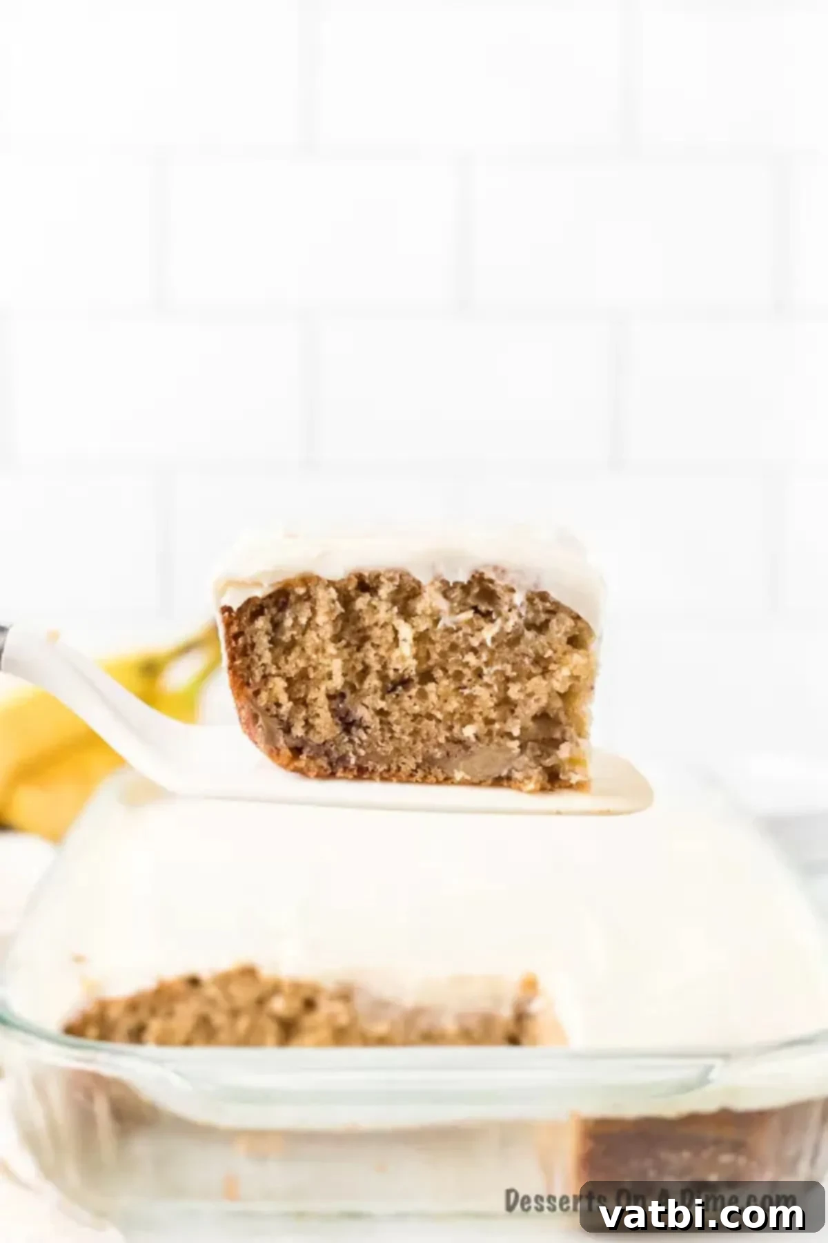 A beautifully frosted banana cake in a baking pan, with one slice served on a spatula, showcasing its moist texture.