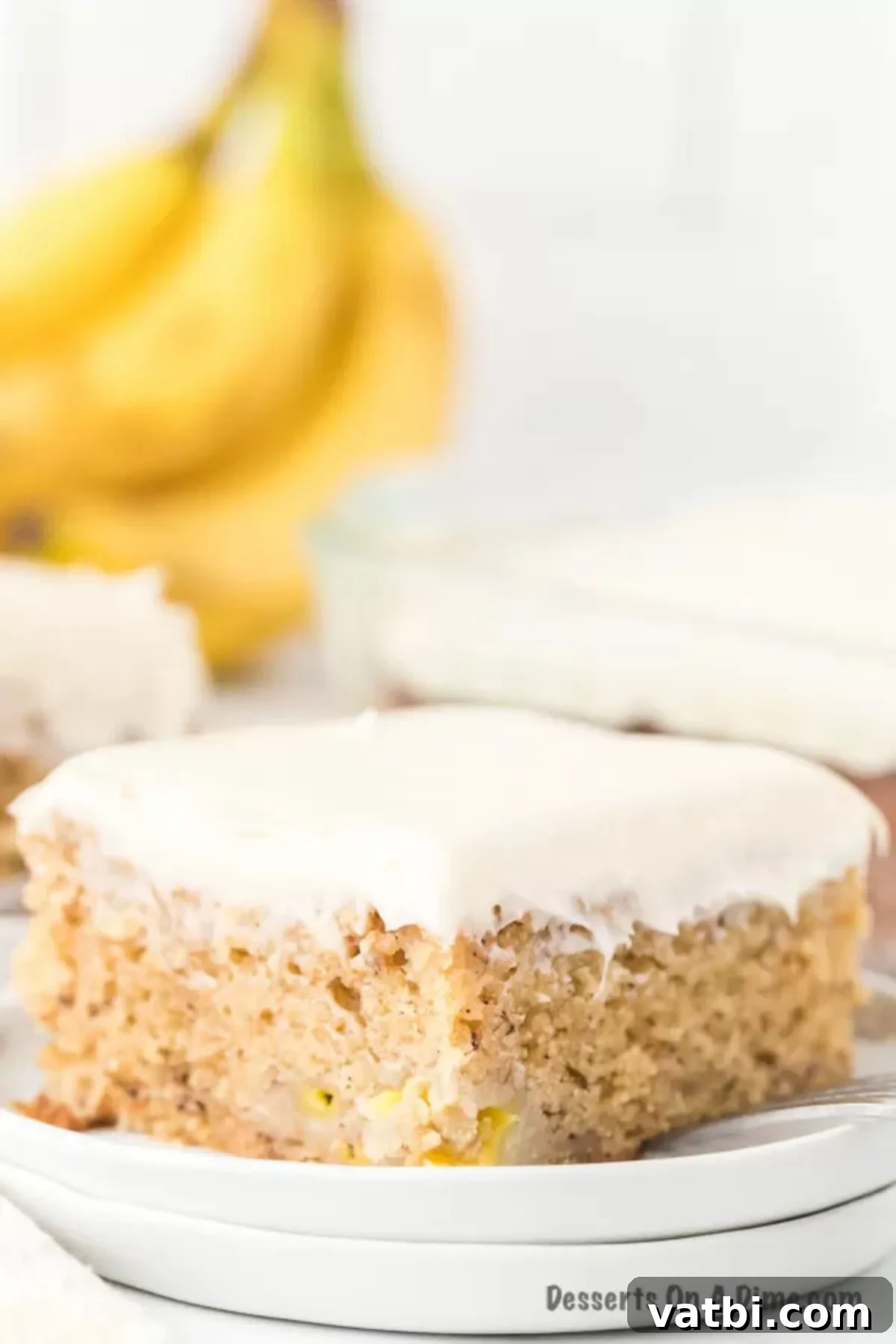 A perfectly sliced piece of moist banana cake topped with creamy cream cheese frosting, resting on a white plate, ready to be enjoyed.