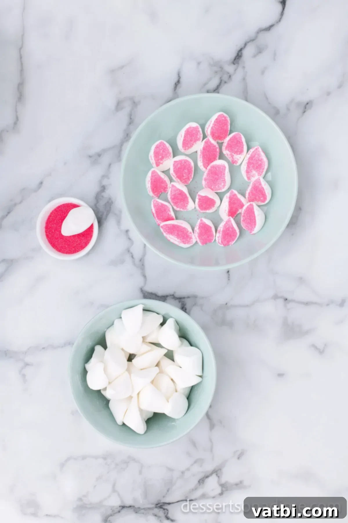 Marshmallows cut diagonally into ear shapes and some dipped in pink sprinkles on a white plate.