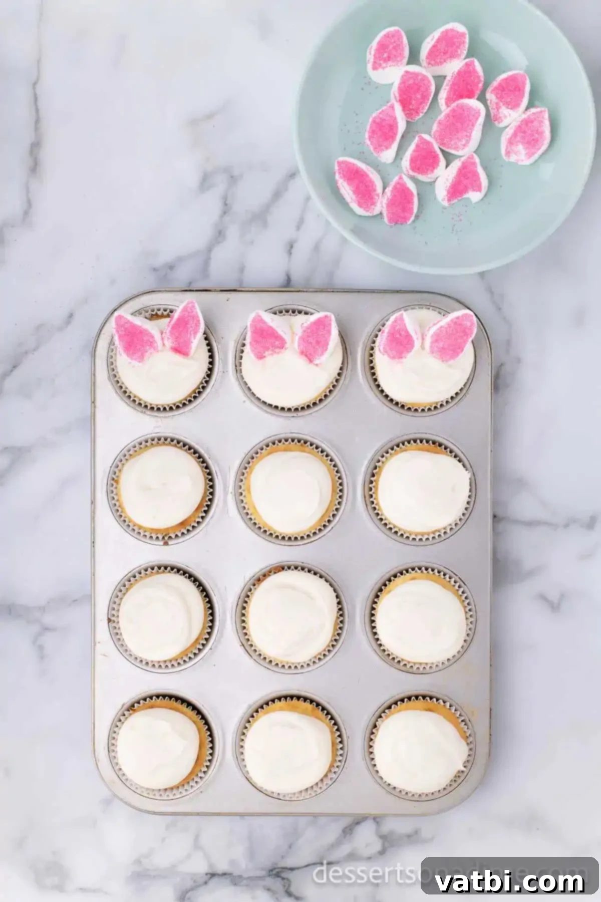 Cupcakes frosted with white icing and topped with pink-sprinkled marshmallow bunny ears, ready for the final touches.