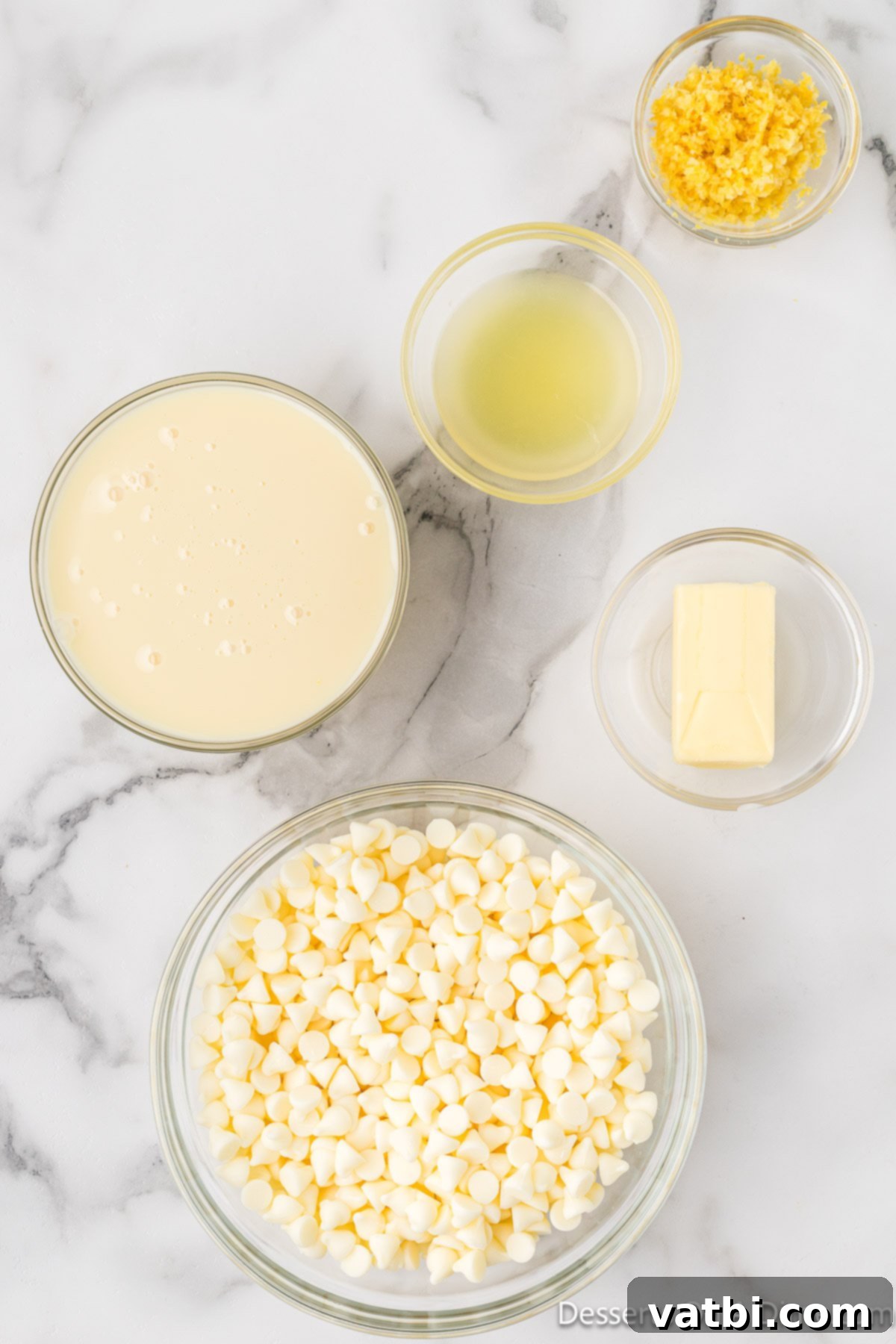 All 5 ingredients for Lemon Fudge: White chocolate chips, sweetened condensed milk, unsalted butter, fresh lemon juice, and lemon zest, laid out on a white surface. 