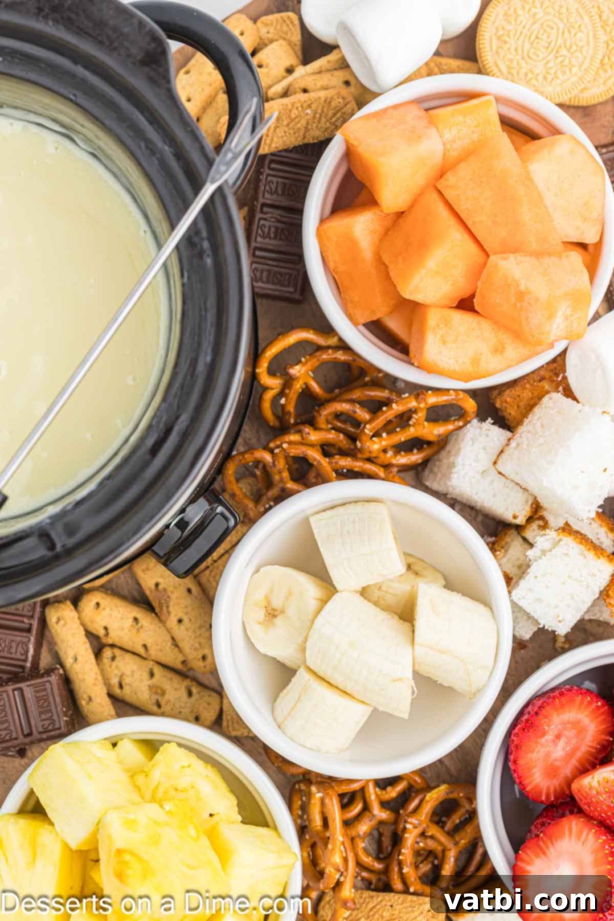 Decadent White Chocolate Fondue 7 White chocolate fondue in a pot surrounded by various fruit and sweet dippers on a table.