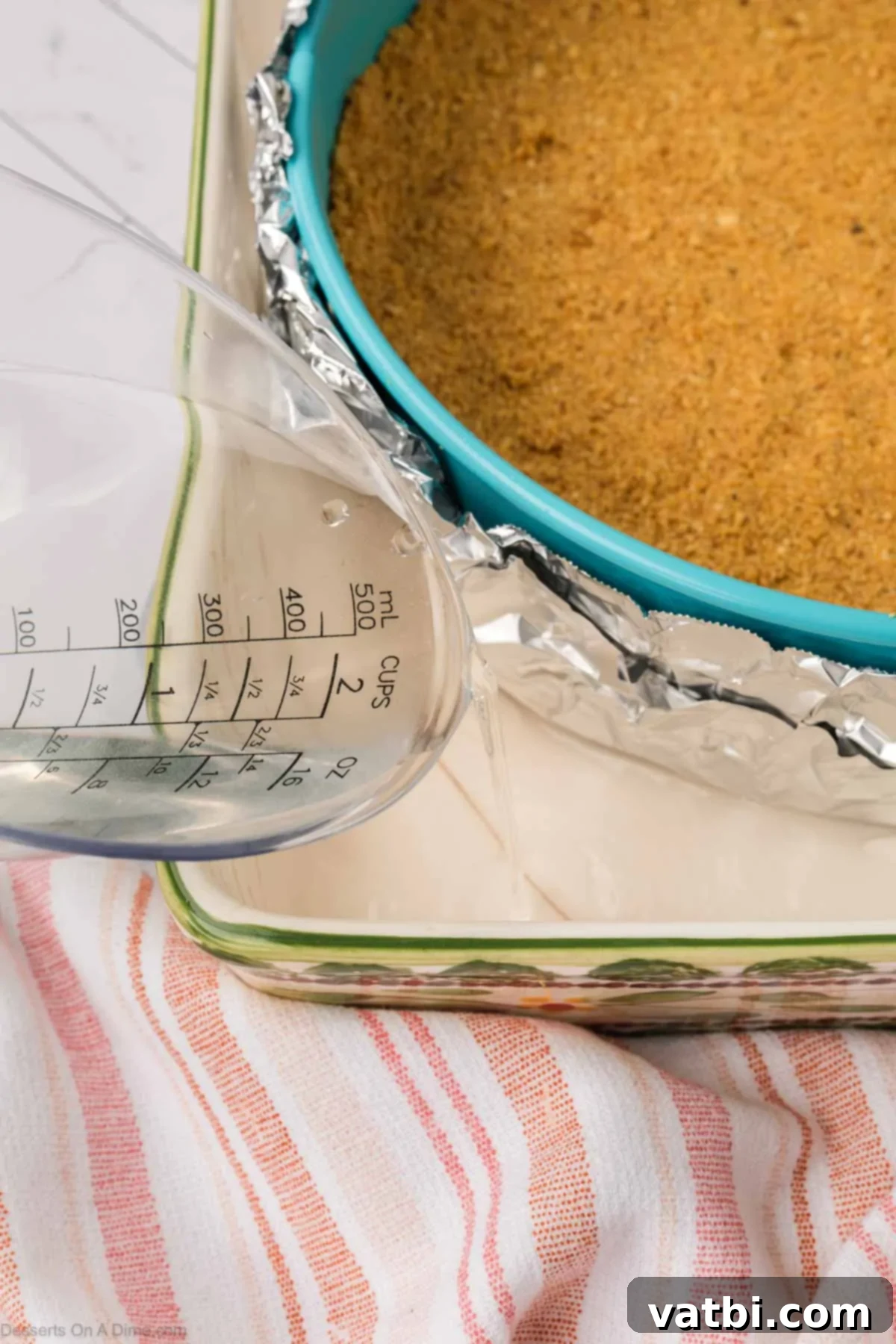 Hot water being carefully poured into a roasting pan around the foil-wrapped cheesecake pan, creating a water bath.